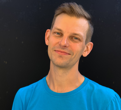 A portrait of Per Nilsson wearing a blue t-shirt and smiling. He has blue eyes, medium-blonde hair, and needs a shave. The background is black.